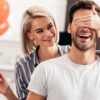mulher atrás de um homem e com a mão nos olhos dele enquanto ela segura um coração e ao fundo balões de coração ao desfoque