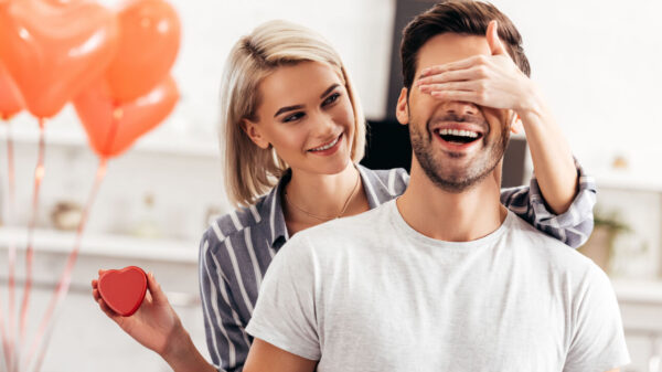 mulher atrás de um homem e com a mão nos olhos dele enquanto ela segura um coração e ao fundo balões de coração ao desfoque