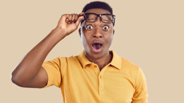 Homem usando uma camisa pólo, com a boca aberta, levantando os óculos, olhando para algo com uma expressão facial surpresa e chocada, em um estúdio.