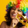 mulher sorrindo segurando uma mascara dourada de carnaval e uma peruca colorida em fundo amarelo