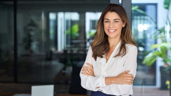 mulher de braços cruzados, usando social e sorrindo em um fundo que parece uma empresa