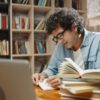 homem sentado em uma biblioteca lendo um livro e com outra pilha de livros ao lado