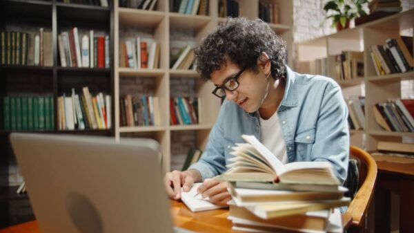 homem sentado em uma biblioteca lendo um livro e com outra pilha de livros ao lado