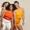mais à esquerda da imagem há três mulheres negras com roupa de academia em um fundo acinzentado