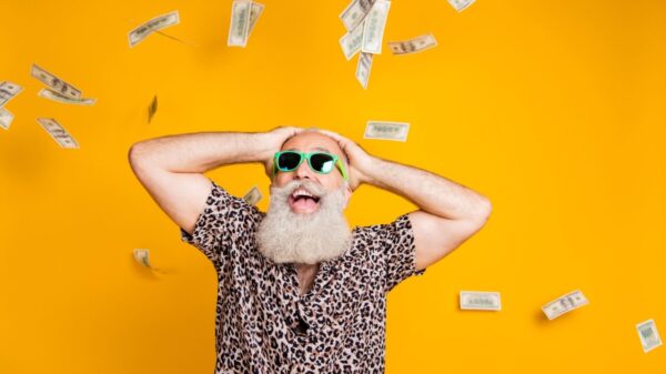 homem com barba grande e branca usando camisa com estampa de onça e com as mãos na cabeça com dinheiro caindo em fundo amarelo