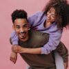 uma mulher negra está nas costas de um homem negro e os dois sorriem em fundo rosa