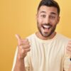homem fazendo o sinal de positivo com as duas mãos e sorrindo em fundo amarelo