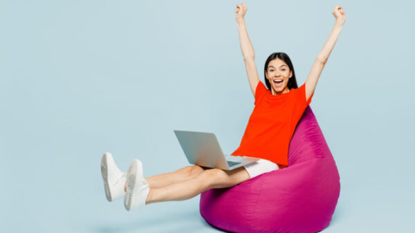 Jovem de corpo inteiro, usando uma camiseta vermelha casual, sentada em um pufe, usando um laptop, fazendo gesto de vencedor, isolada em um estúdio de fundo azul simples. Conceito de estilo de vida.
