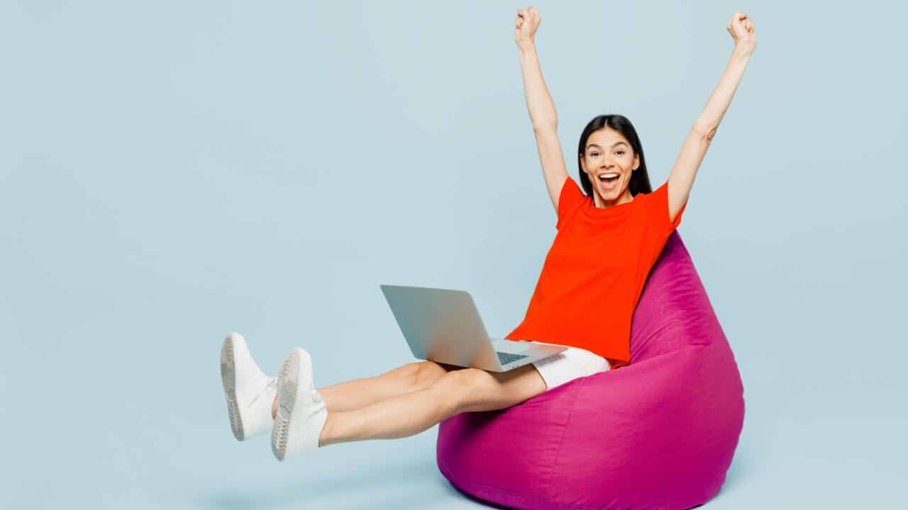 Jovem de corpo inteiro, usando uma camiseta vermelha casual, sentada em um pufe, usando um laptop, fazendo gesto de vencedor, isolada em um estúdio de fundo azul simples. Conceito de estilo de vida.