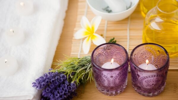 à esquerda tá parte de uma banheira branca e quadrada cheia de espumas e à direta há velas lavandas e margaridas em cima de uma mesa de madeira