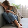 mulher sentada em um sofá cinza claro com um celular na mão ela esconde o rosto entre o braço