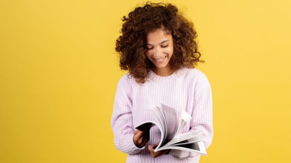 Mulher feliz lendo um livro, passando as páginas, sorrindo, de pé sobre um fundo amarelo.