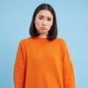 mulher ao centro com cara chateada vestindo moletom laranja em fundo azul claro