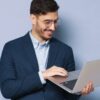No centro da imagem está um homem com roupa social azul segurando um notebook cinza em um fundo roxo