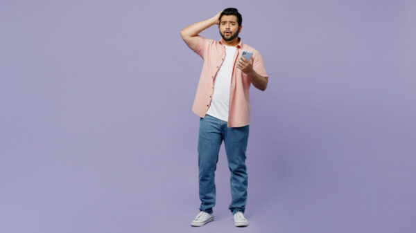 Homem chocado, assustado, usando uma camisa rosa e uma camiseta branca por baixo, roupas casuais, segurando um celular, com a mão na cabeça, isolado em um estúdio de fundo roxo pastel simples.