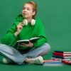 Retrato de uma jovem estudante sentada no chão uma com expressão pensativa, estudando, isolada sobre um fundo verde de um estúdio. Conceito de educação, estudo, lição de casa, juventude, estilo de vida.