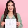 Foto de uma mulher mostrando um calendário, olhando para o lado, usando uma roupa verde, isolada em um fundo de cor rosa pastel.