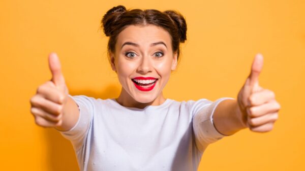 mulher usando roupa branca e batom vermelho, sorrindo e fazendo sinal de joia com as duas mãos em fundo amarelo