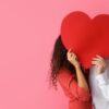 casal no canto direito com imagem se beijando com os rostos escandidos com um grande coração vermelho de cartolina em fundo rosa