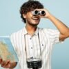 homem olhando com binóculos e uma câmera fotográfica no pescoço em fundo azul