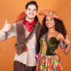 casal com roupas típicas de festa junina abraçados e fazendo o sinal de joia com a mão em fundo laranja