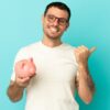 homem jovem com camisa branca segurando um porquinho de cofre em fundo azul claro