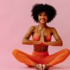 mulher com roupa salmão de ioga e em posição de meditação em fundo rosa