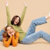 Duas mulheres felizes de corpo inteiro, uma usando camisa casual verde e, outra, laranja, uma sentada e, outra, deitada. Uma faz um gesto de vencedora. Isoladas em um fundo bege pastel liso. Conceito de estilo de vida.