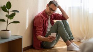 homem sentado no chão com expressão de nervosismo e segurando um celular