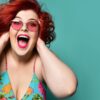 mulher de cabelo e óculos vermelhos com a boca aberta de felicidade em fundo verde água