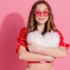 menina com óculos vermelho em formato de coração e abraçada com uma almofada de coração em fundo rosa