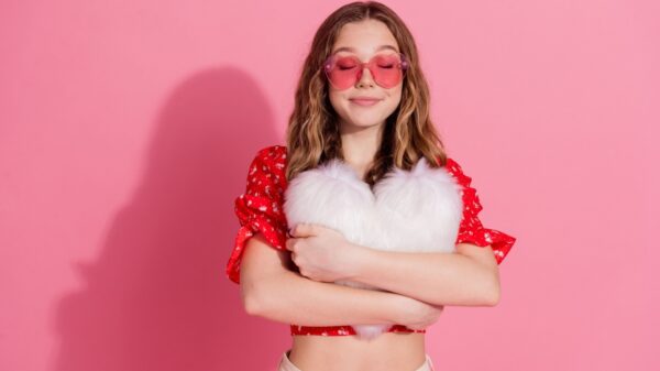 menina com óculos vermelho em formato de coração e abraçada com uma almofada de coração em fundo rosa