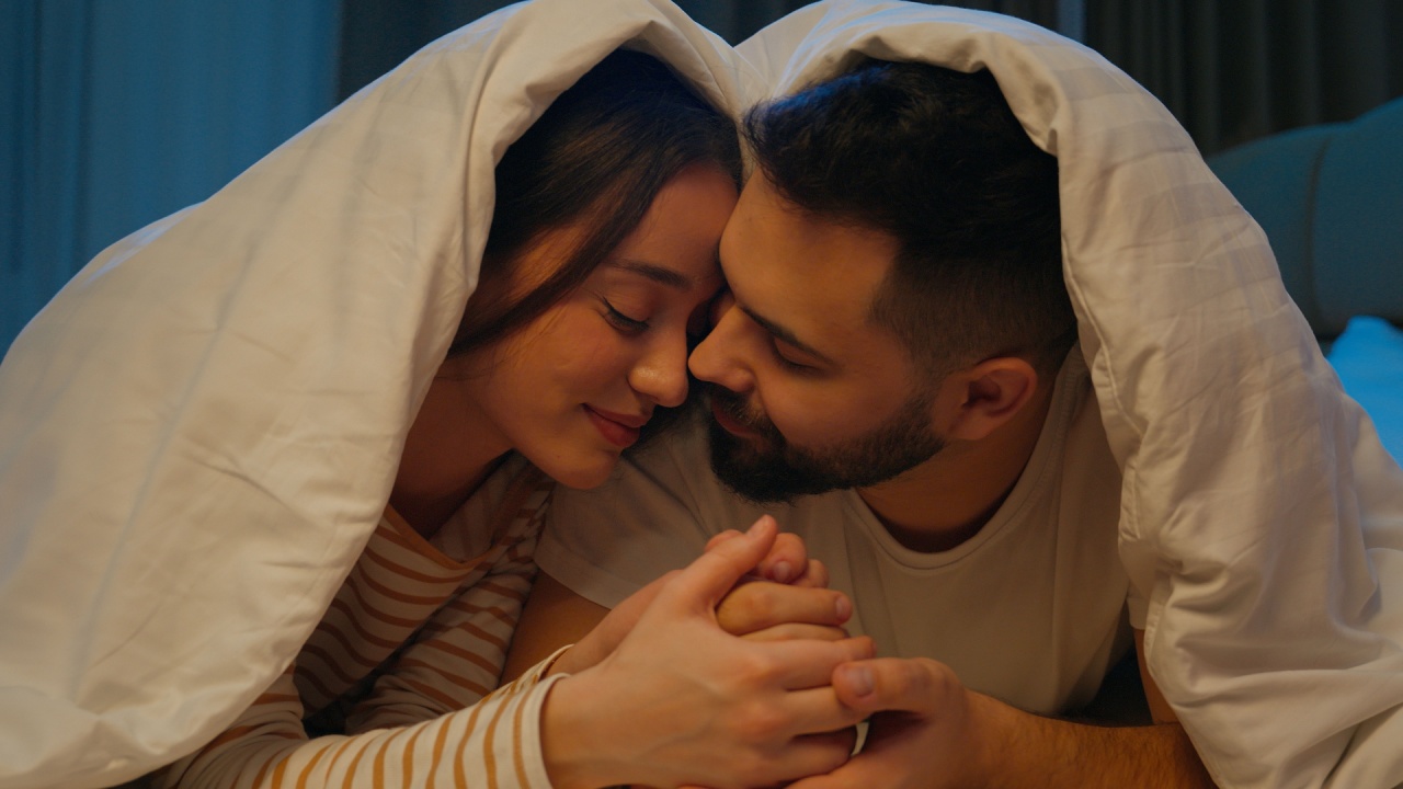casal deixados na cama e com edredom por cima com as mãos dadas e sorrindo