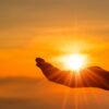 silhueta de uma mão posicionada como se estivesse segurando o sol em um céu alaranjado