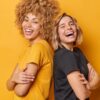 duas mulheres rindo e encostadas nas costas uma da outra em fundo amarelo