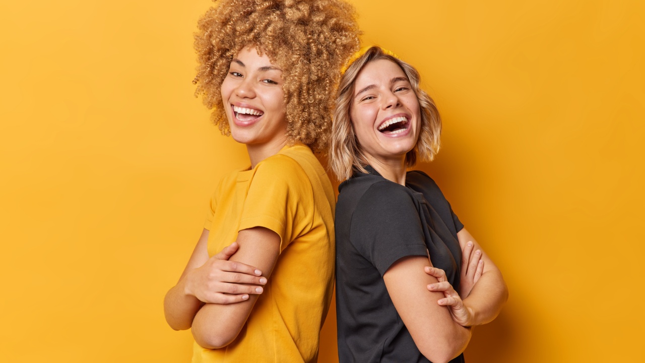 duas mulheres rindo e encostadas nas costas uma da outra em fundo amarelo