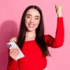 Mulher sorrindo feliz e comemorando com um celular na mão direita e em fundo rosa