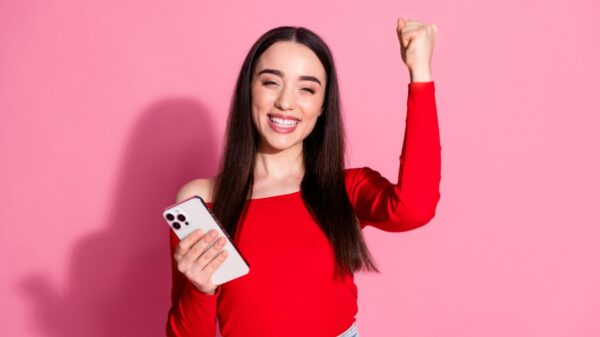 Mulher sorrindo feliz e comemorando com um celular na mão direita e em fundo rosa