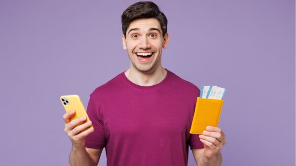 Homem usando roupas casuais, segurando bilhetes de passagem aérea e um celular isolado em um fundo roxo pastel plano. Conceito de viagem aérea.