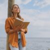 mulher encostada em um poste lendo um livro ao ar livre e próxima ao mar