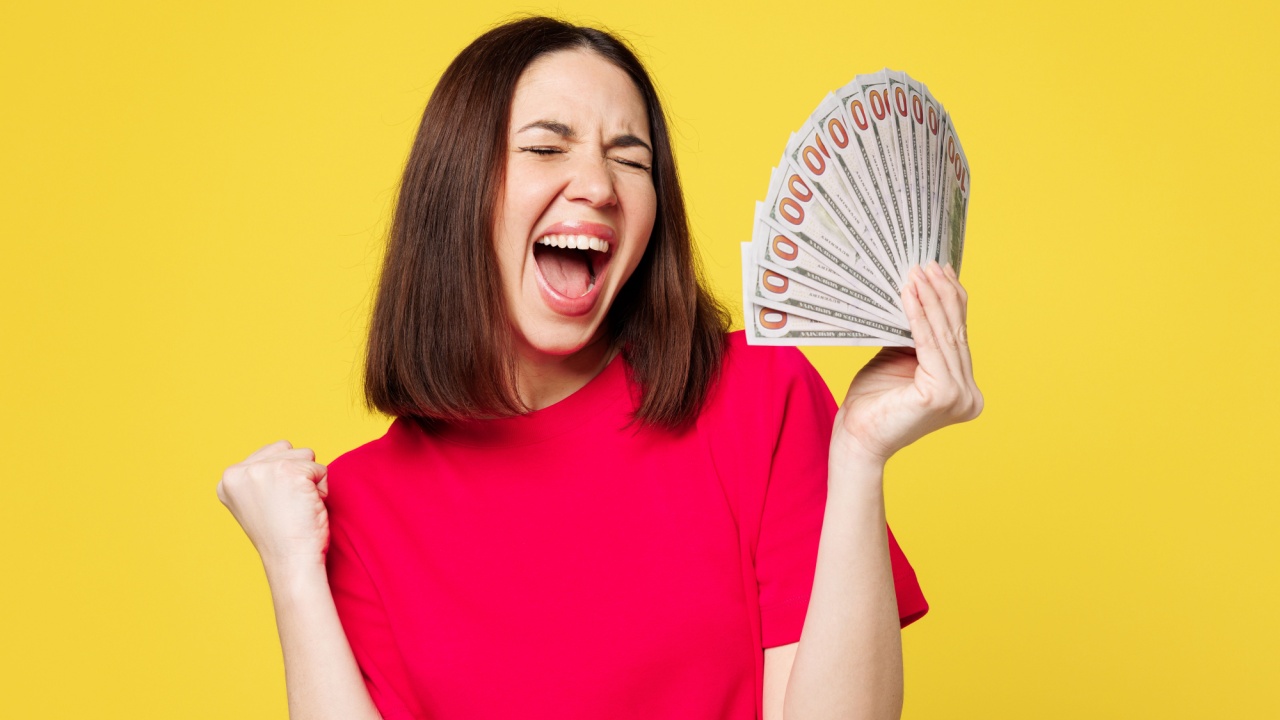mulher segurando um leque de dinheiro e com expressão facial de comemoração com a boca aberta e em fundo amarelo