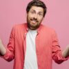 homem de roupa branca e rosa segurando um celular quebrado na mão direita em fundo rosa
