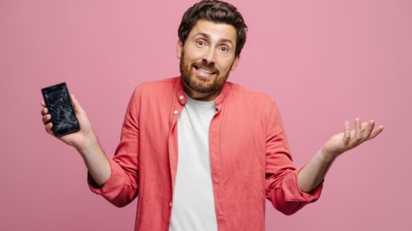 homem de roupa branca e rosa segurando um celular quebrado na mão direita em fundo rosa