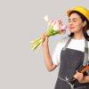 mulher com roupa de operária segurando um martelo em uma mão e um buquê de rosas na outra mão e na altura do nariz em fundo cinza