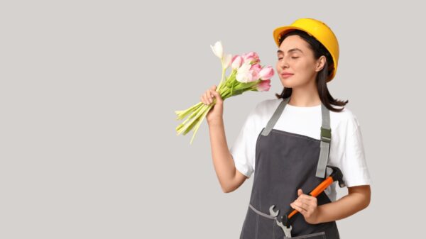 mulher com roupa de operária segurando um martelo em uma mão e um buquê de rosas na outra mão e na altura do nariz em fundo cinza