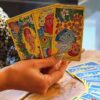 imagem de uma mão feminina segurando três cartas de tarot e um deck de cartas em uma mesa de madeira