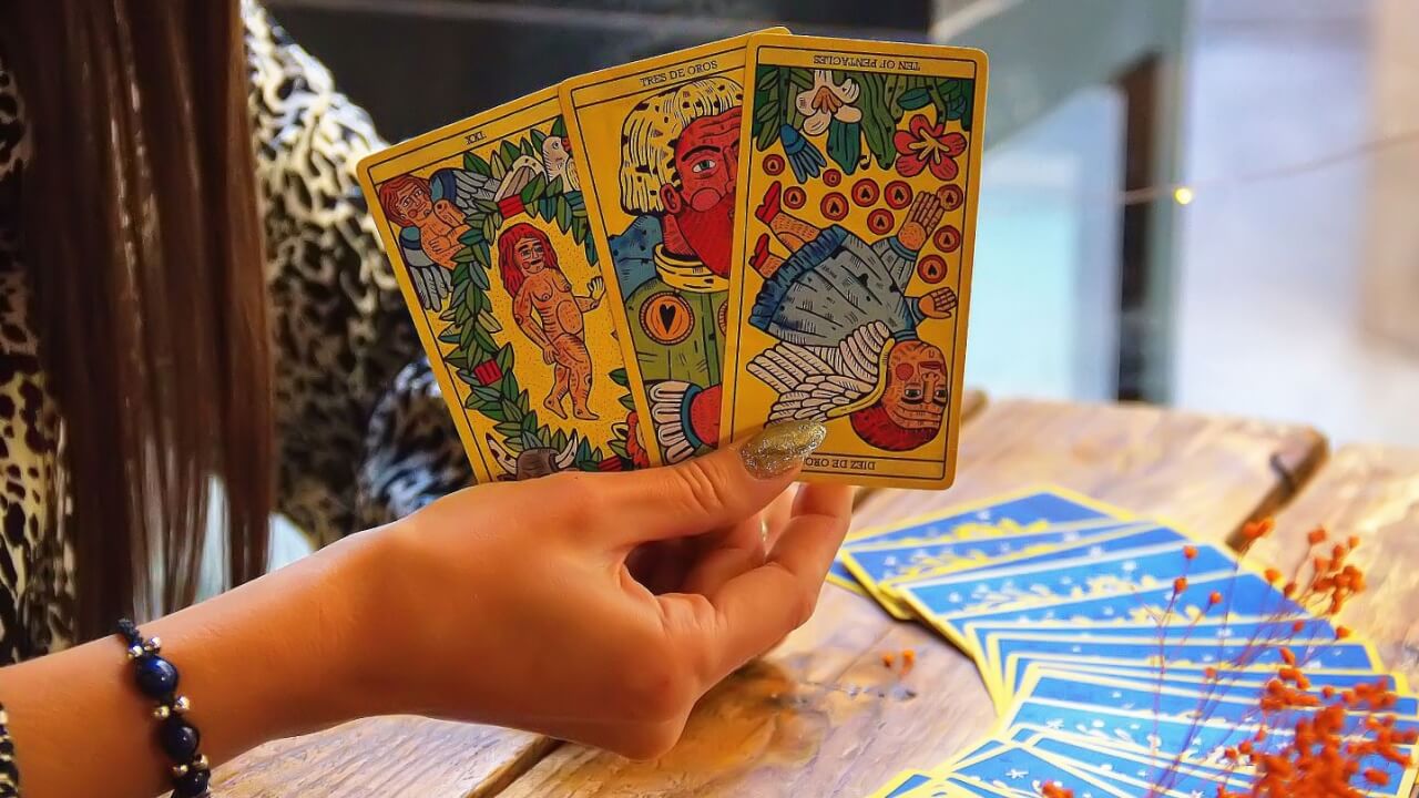 imagem de uma mão feminina segurando três cartas de tarot e um deck de cartas em uma mesa de madeira