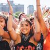 três mulheres jovens na grade do festival Lollapalooza com as mãos no alto