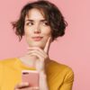 mulher com o dedo indicador no queixo olhando para o lado e pensando e segurando um celular em fundo rosa