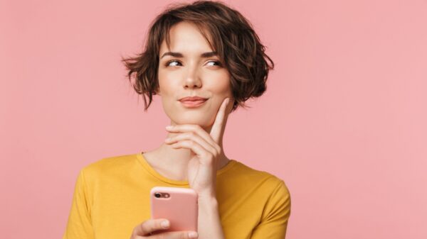 mulher com o dedo indicador no queixo olhando para o lado e pensando e segurando um celular em fundo rosa